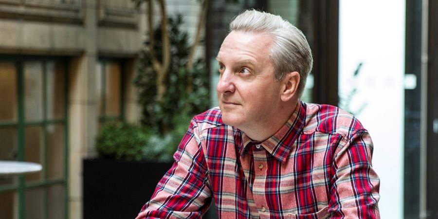 A man with short, graying hair wearing a red and blue plaid shirt is looking off to the side.