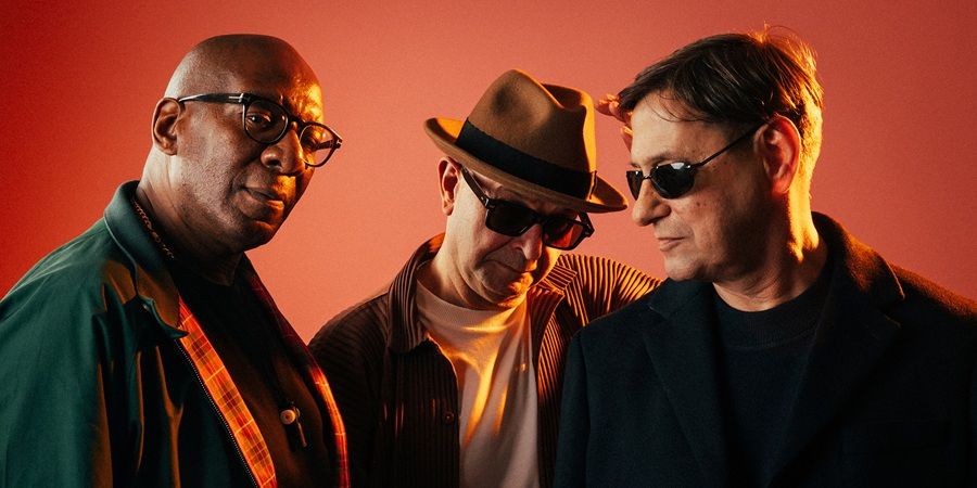 Three men posing for a photo against a red background. The man on the left wears glasses and a green jacket. The middle man wears a brown fedora and sunglasses. The man on the right wears sunglasses and a black jacket.