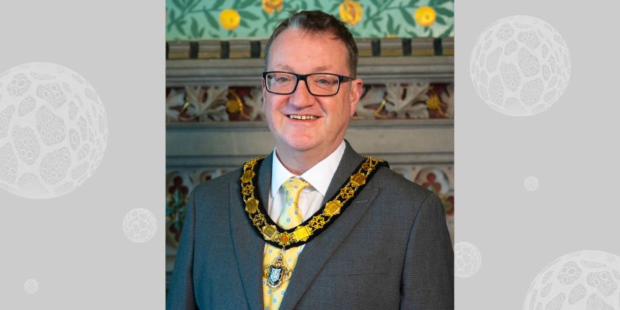 A man wearing a suit, a yellow tie with blue dots, and a ceremonial chain with a pendant.