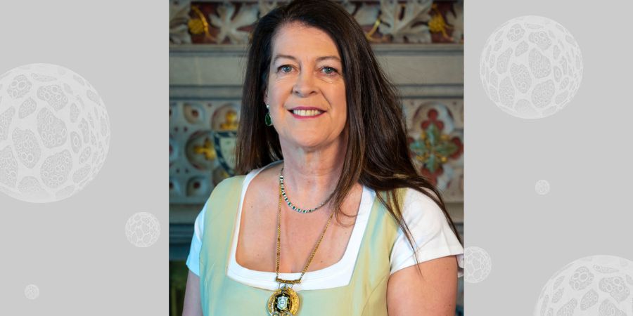 A person wearing a light green dress over a white shirt, adorned with a gold medal and a green beaded necklace. The background features ornate architectural details.