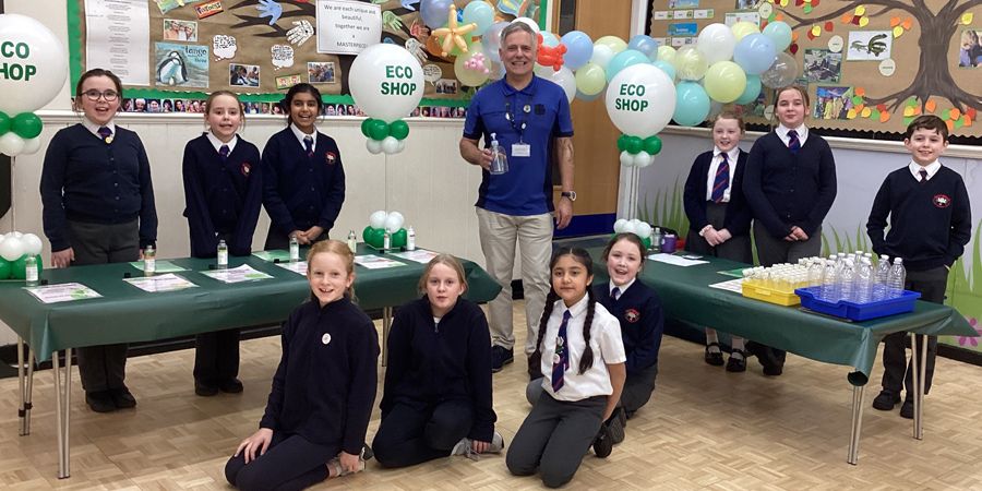 A group of children in school uniforms stand behind tables at an Eco Shop, with balloons and posters in the background. 
