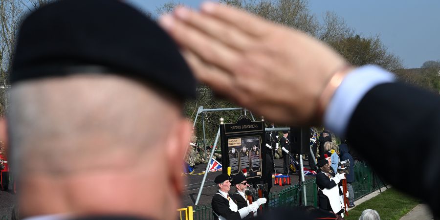 A person in a black beret salutes during an outdoor ceremony with other uniformed individuals in the background.