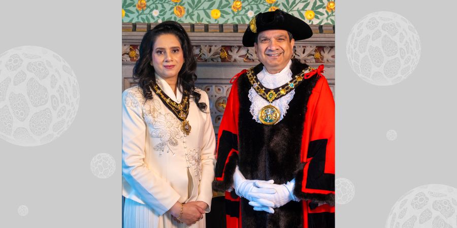 A woman in a cream-colored formal dress with a ceremonial necklace stands beside a man in a red and black ceremonial robe, white ruff, and mayoral chain.