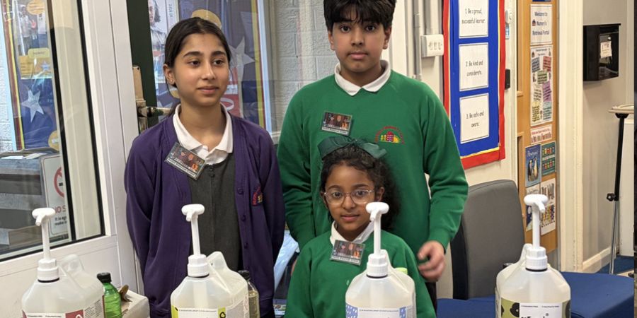 3 children wearing school uniforms stand in front of large pump bottles.