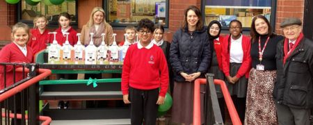 A group of people, including students in red uniforms and adults, stand in front of a display of bottles on a tiered stand outside a building.