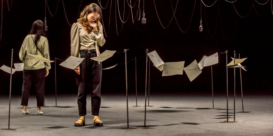 A person stood in a a dark room, surrounded by cables and microphones suspended from the ceiling.