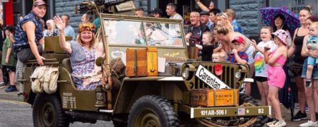 A vintage military jeep with people dressed in 1940s attire drives through a street parade, with onlookers watching from the pavement.