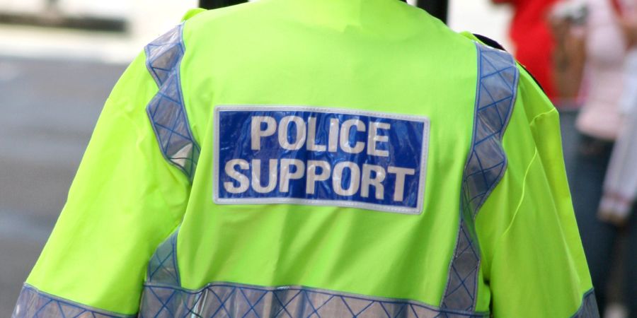 A person in a bright yellow police support jacket and a black hat walks down a street.