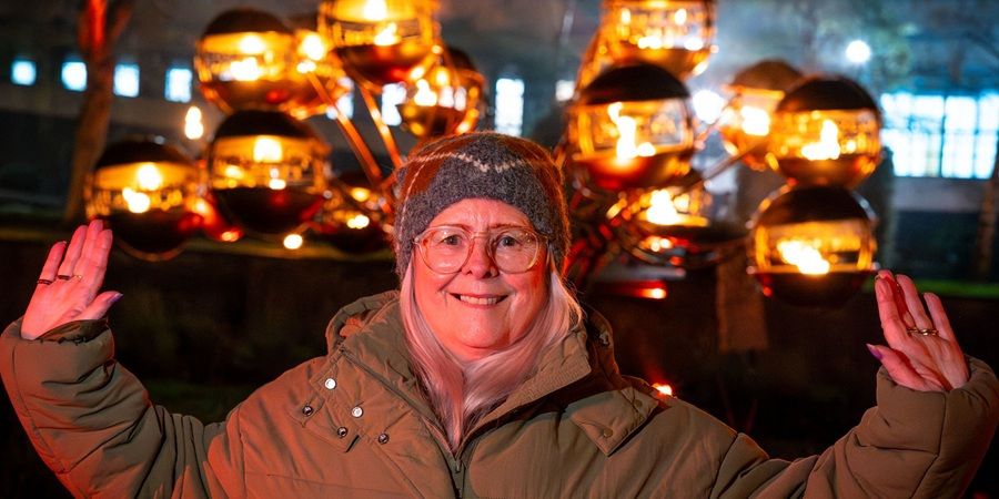 A person in a winter coat and hat smiles at the camera with arms outstretched, standing in front of a display of glowing, orb-shaped lanterns.
