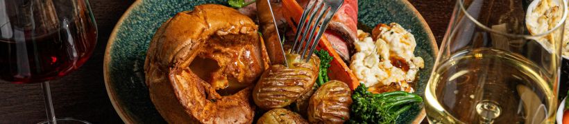 A plate of roast beef with Yorkshire pudding, roasted potatoes, broccoli, carrots, and cauliflower cheese, served with glasses of red and white wine.