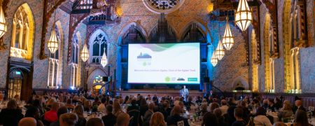 A speaker stands on a stage in front of a large screen in a grand hall filled with people seated at round tables for a formal event.