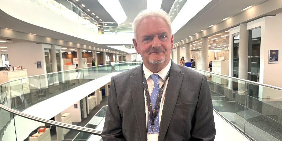 A man in a dark suit and blue patterned tie stands in a modern office atrium.