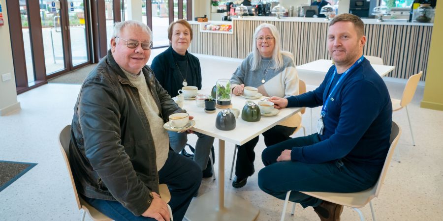 4 people sitting around a table with cups and teapots, with a cafe counter in the background.
