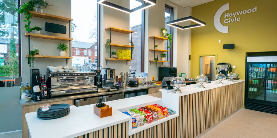A modern cafe counter with a professional espresso machine, shelves with plants, and a refrigerator stocked with drinks. The "Heywood Civic" logo is visible on the wall.