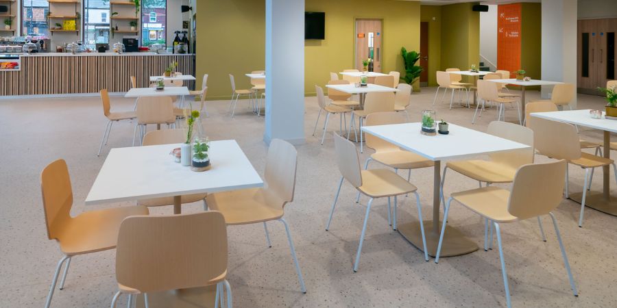 A modern cafe interior with many white tables and light wood chairs, a counter with coffee machines and shelving in the background, and large windows letting in natural light.