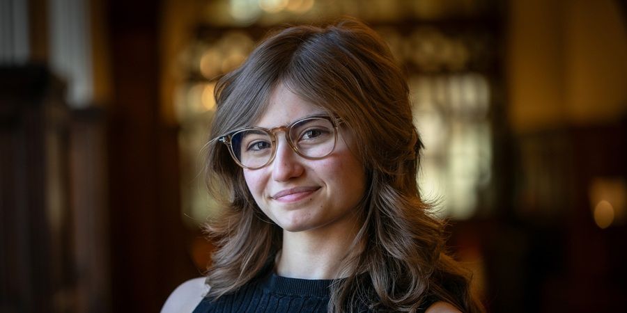 A person with long, wavy brown hair and glasses smiles gently.