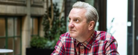 A person wearing a red and white plaid shirt looks off to the side, with a blurred background of plants and architecture.
