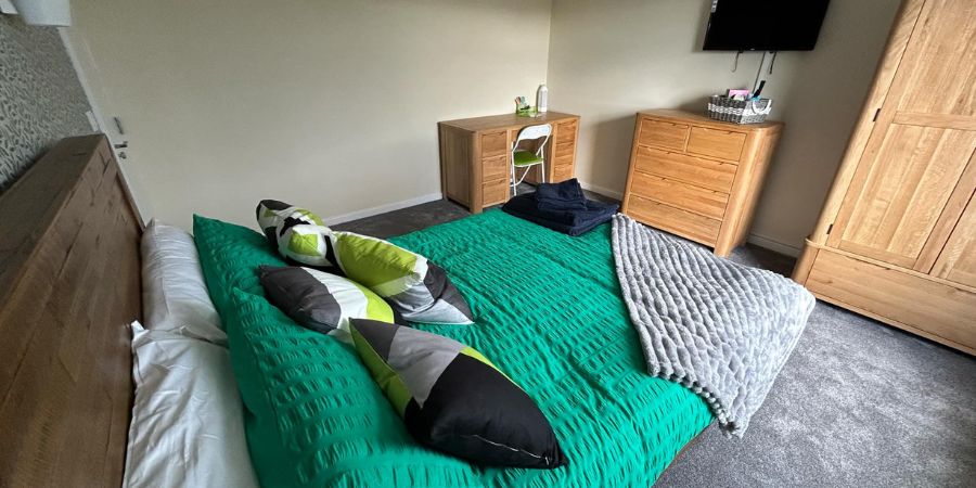 A bedroom with a wooden bed frame, a green blanket, and decorative pillows. There is also a wooden dresser, a desk with a chair, and a large wardrobe.