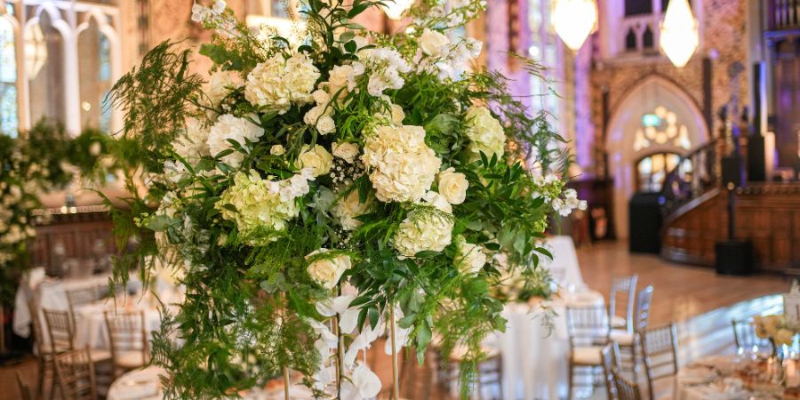 A tall, elaborate floral arrangement of white hydrangeas and roses with lush greenery sits in the foreground, with a blurred background of a banquet hall featuring tables, chairs, and ornate architecture.