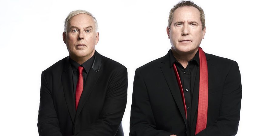 Two men in black suits and red ties stand against a white background.