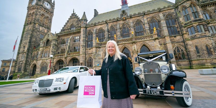 A person holds a white shopping bag with "Rochdale Wedding Show" printed on it, standing between a white modern car and a vintage car in front of a large, ornate building.