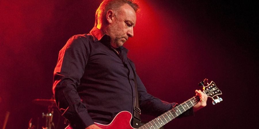 A man with short hair and a beard plays a red electric guitar on stage with red lighting.