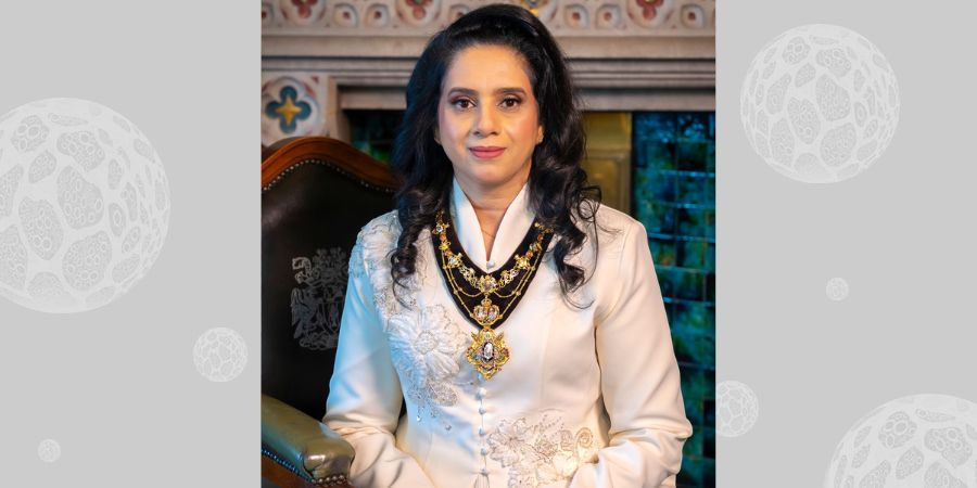 A woman in a white formal outfit and elaborate necklace sits in a chair, with a decorated fireplace behind her.