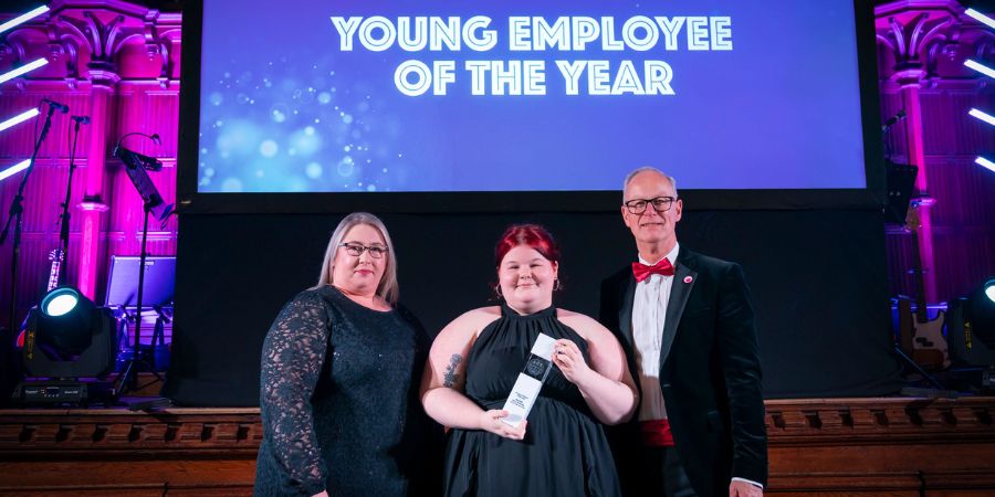 3 people stand on stage holding an award in front of a backdrop reading "Young Employee of the Year".