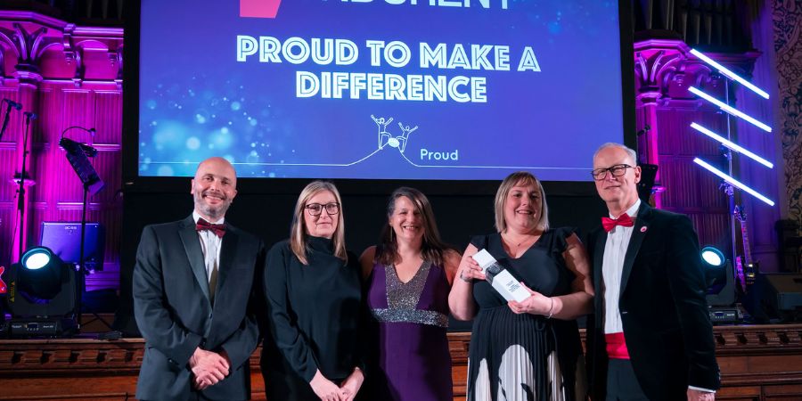 A group of 5 people standing on stage in formal attire, holding an award, with a backdrop featuring the words "Proud to Make a Difference."