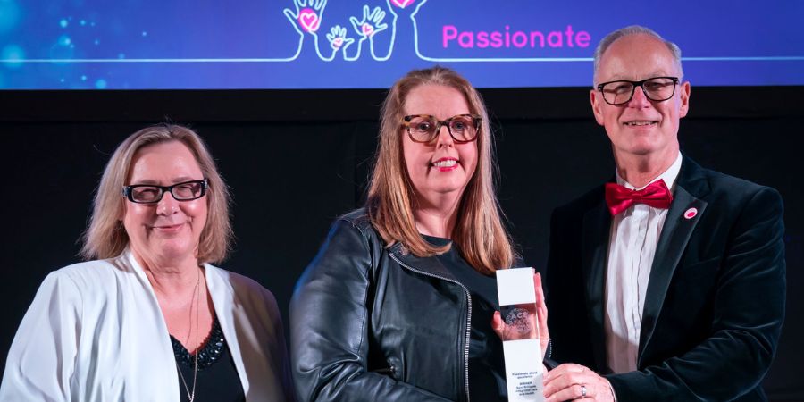 3 individuals stand together on stage, smiling and holding an award, against a colourful backdrop.