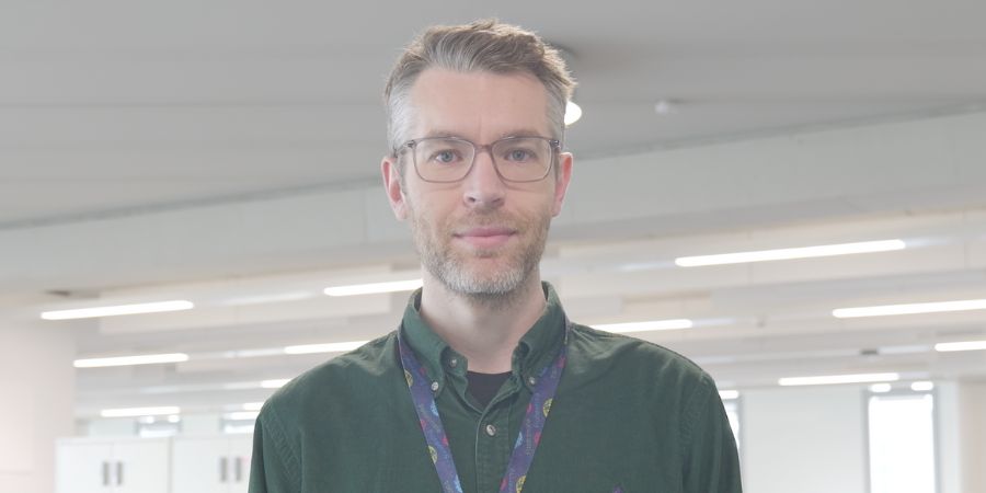 A man in a green shirt and lanyard stands in a modern office space, with desks and lockers visible in the background.