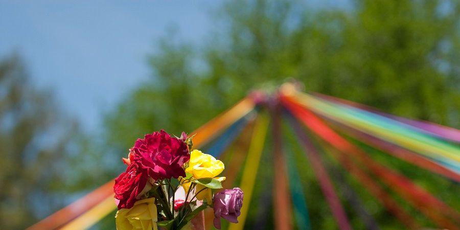 A vibrant bouquet of red, yellow, and pink roses in focus against a blurred background of a maypole with colourful ribbons under a bright blue sky.