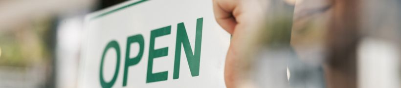 A hand places an "Open" sign on a glass door, viewed from the outside with reflections.