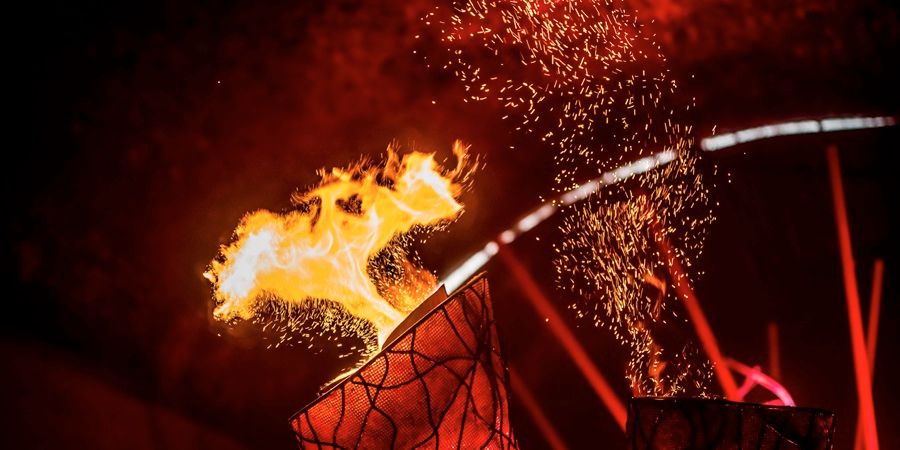 A fiery flame bursts from a burning hoop-shaped structure as red spotlights shine in the background, with sparks and glowing embers flying through the air.