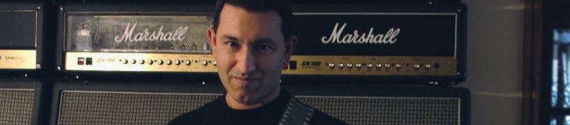 Aziz Ibrahim holding his guitar, stood in front of several amplifiers.