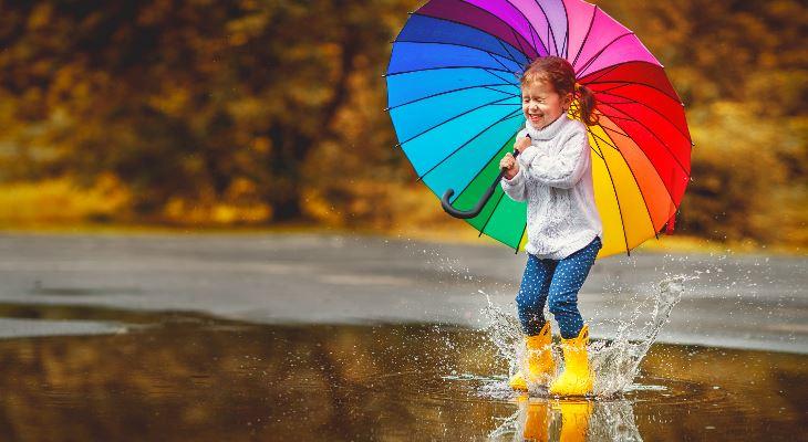 A child with a rainbow umbrella joyfully splashing in a large puddle.