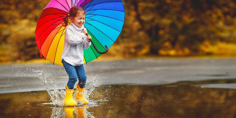 A young girl holding a rainbow umbrella, splashing in a large puddle.