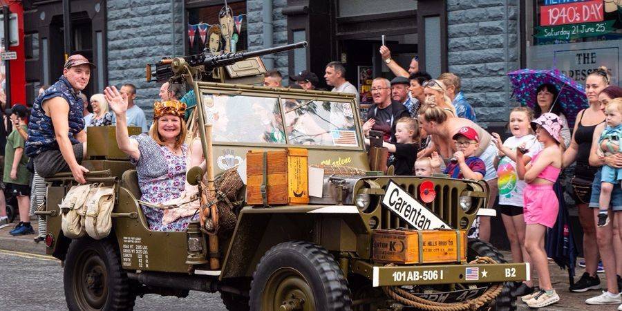 A vintage military jeep with people dressed in 1940s attire drives through a street parade, with onlookers watching from the pavement.