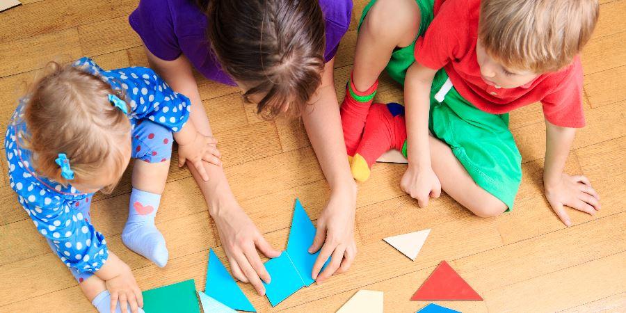 A childminder and children playing with geometric shapes on the floor.