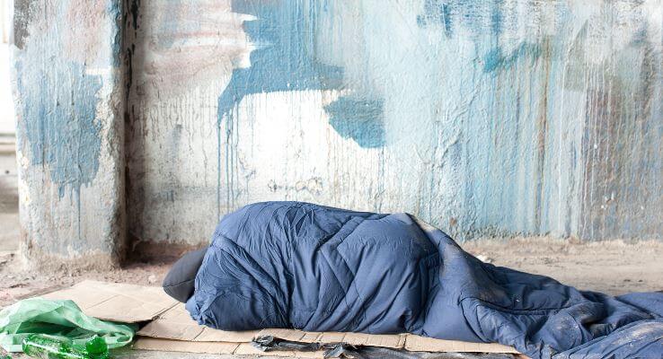 A homeless person in a sleeping bag on cardboard under a bridge.