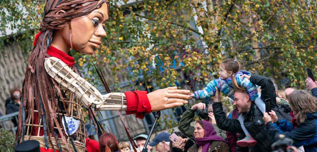 Little Amal reaching out to greet a young girl on her father's shoulders.