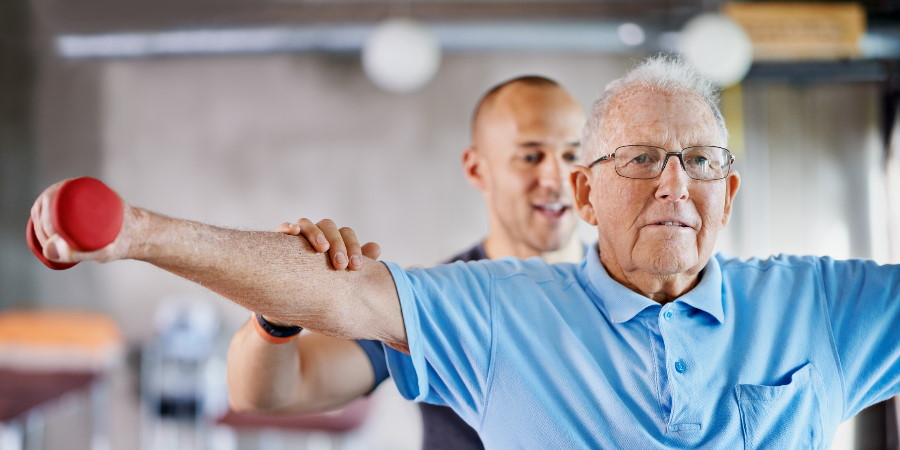 Elderly person exercising, supported by a trainer.