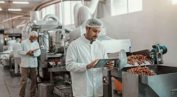 An inspector in a lab coat and hairnet assessing a meat grinding tool.