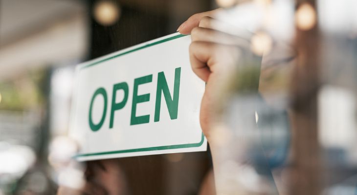 A hand places an "Open" sign on a glass door, viewed from the outside with reflections