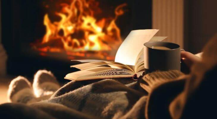 A person reading a book in front of a roaring fireplace.
